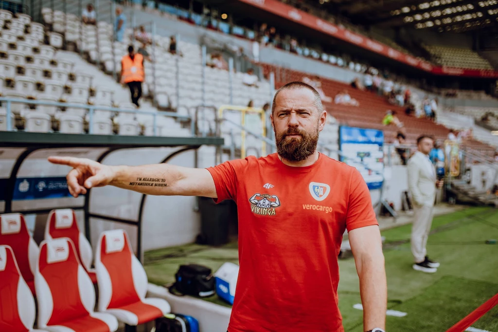 Adam Fudali, kierownik drużyny w ekipie gliwickiego Piasta Mężczyzna z brodą ubrany w czerwoną koszulkę drużyny sportowej stoi na stadionie i wskazuje ręką w lewo, w tle puste krzesełka trybun oraz kilka osób przy murawie.
