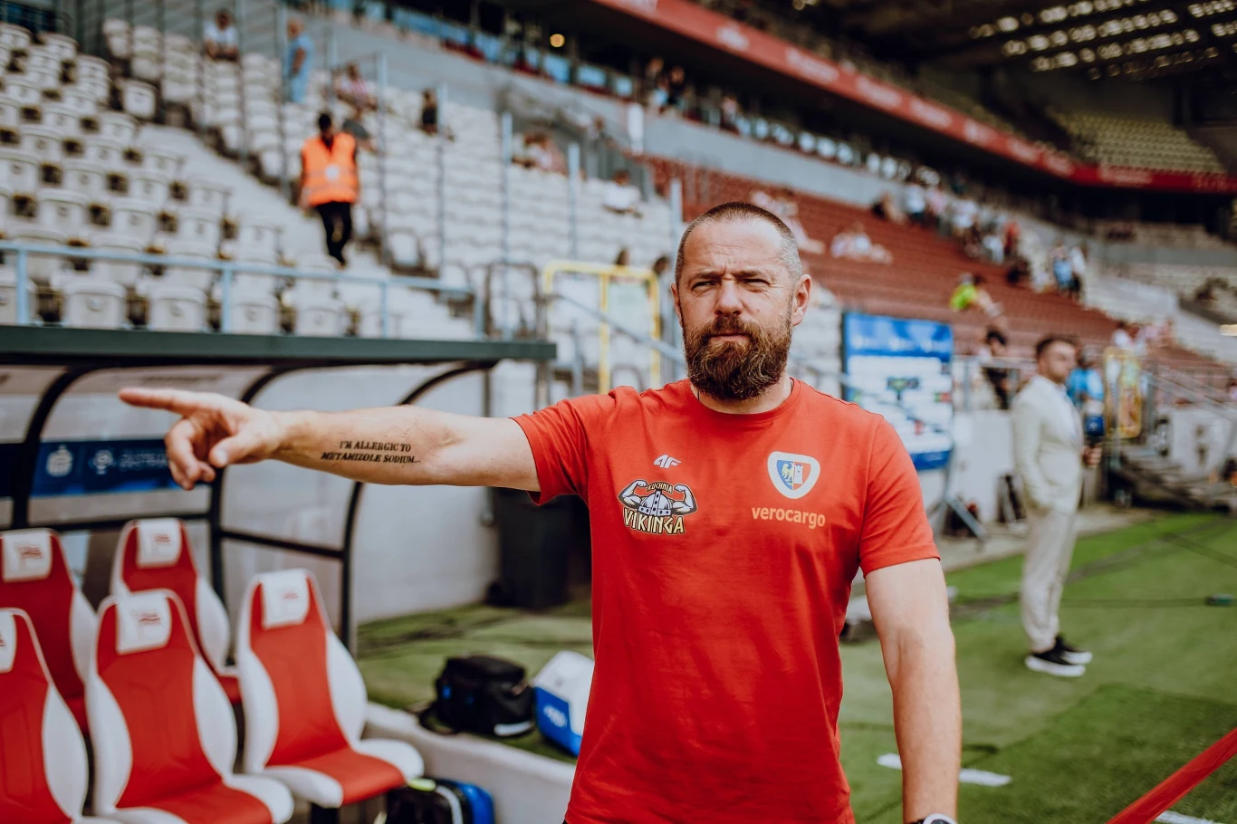 Mężczyzna z brodą ubrany w czerwoną koszulkę drużyny sportowej stoi na stadionie i wskazuje ręką w lewo, w tle puste krzesełka trybun oraz kilka osób przy murawie.