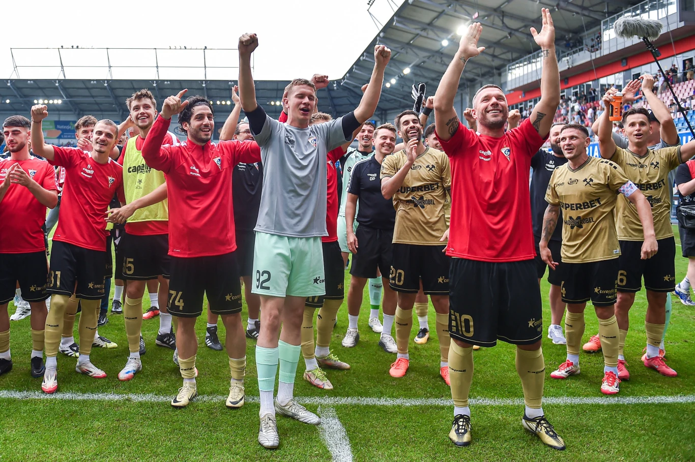 Zawodnicy drużyny piłkarskiej świętują zwycięstwo na murawie stadionu, unosząc ręce i wyrażając radość, wokół nich zgromadzeni są kibice na trybunach.
