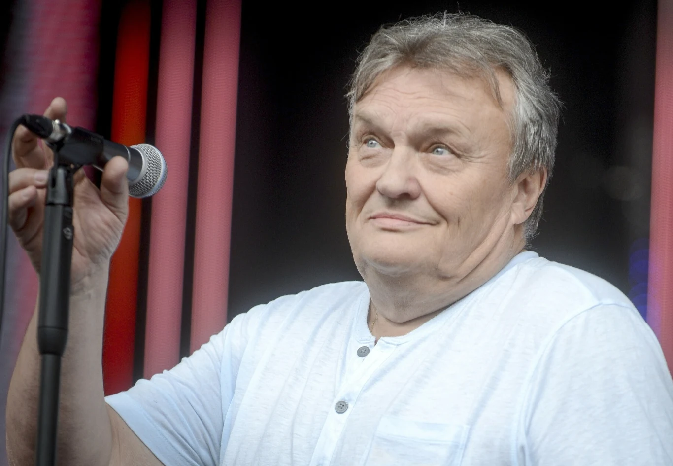Krzysztof Cugowski, wokalista Budki Suflera Mężczyzna w średnim wieku ubrany w jasną koszulkę trzyma mikrofon i patrzy z uśmiechem w bok, na tle ciemnych i różowych pionowych linii.