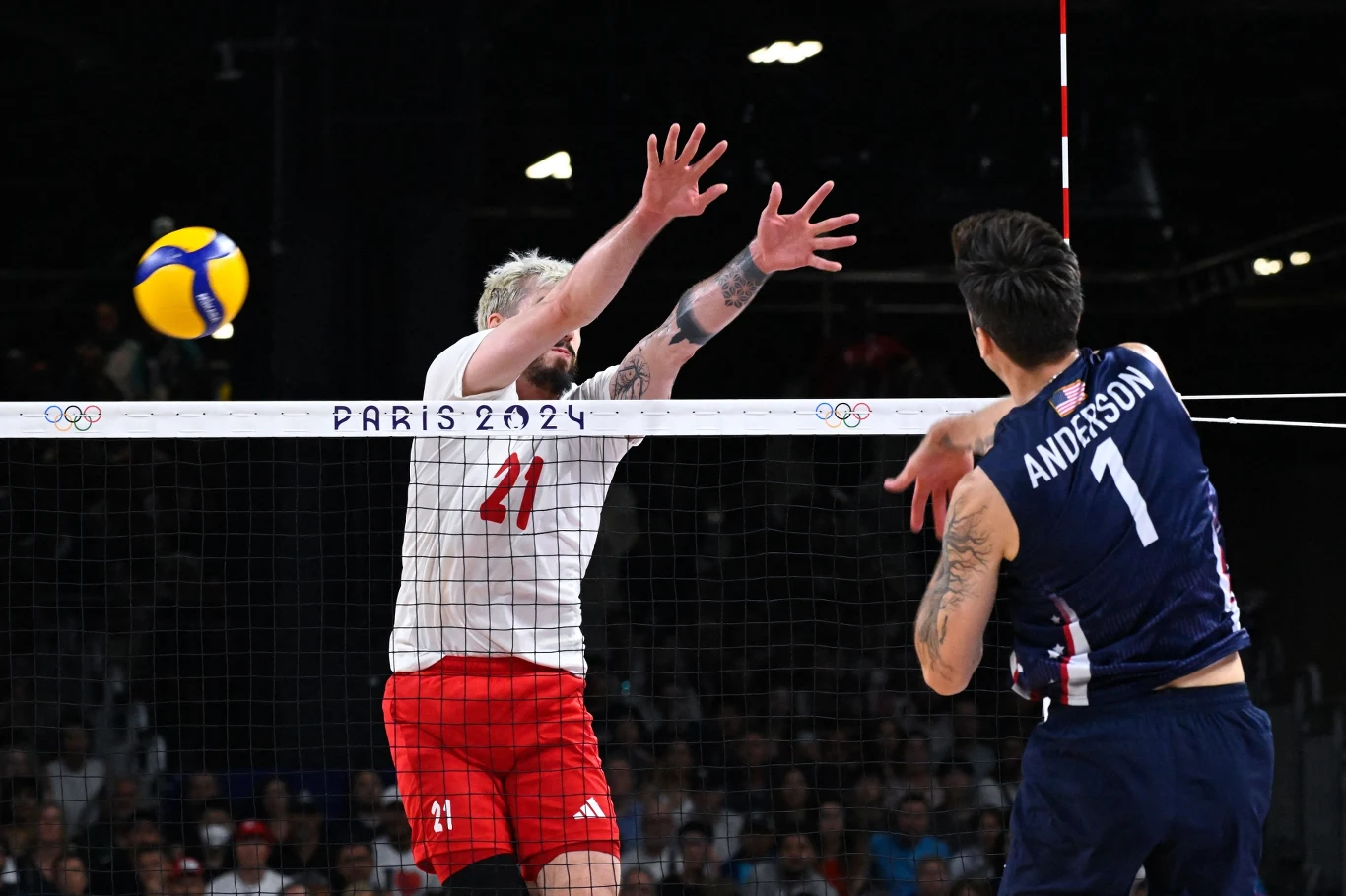 Tomasz Fornal Dwóch siatkarzy walczy przy siatce podczas meczu olimpijskiego, zawodnik w białej koszulce stara się zablokować atakującego w granatowym stroju, nad nimi widoczna piłka oraz napis Paris 2024 na siatce.