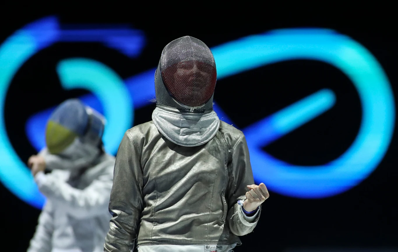 Zuzanna Cieślar Szermierz ubrany w pełny strój ochronny i maskę z sitem, na tle jasnych, neonowych kształtów, wykonuje gest przy użyciu dłoni. W tle widoczna kolejna postać również w stroju szermierskim.