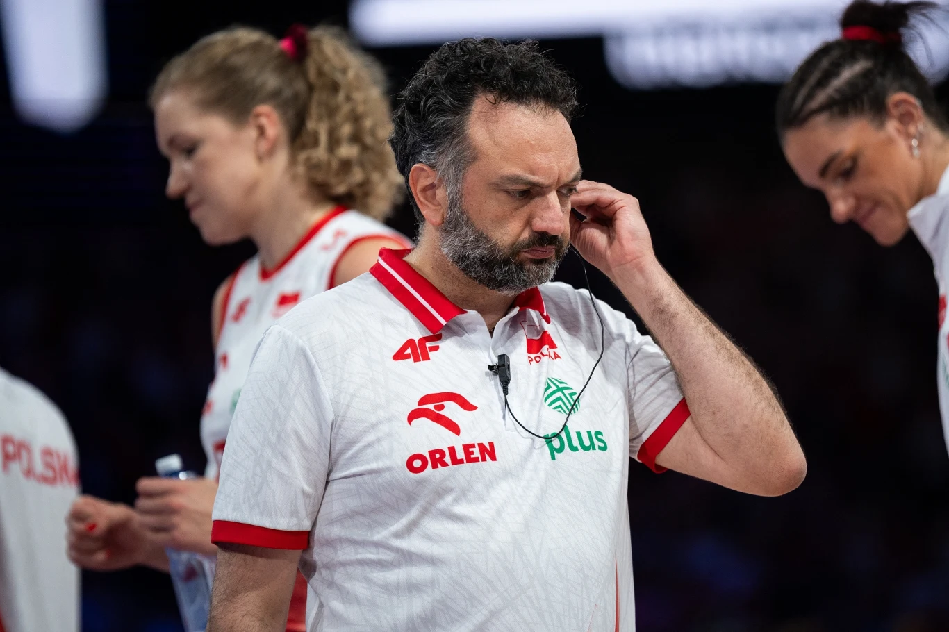 Stefano Lavarini Trener reprezentacji Polski w koszulce z logo sponsorów, w towarzystwie siatkarek w tle, koncentruje się podczas wydarzenia sportowego, sprawiając wrażenie zamyślonego.