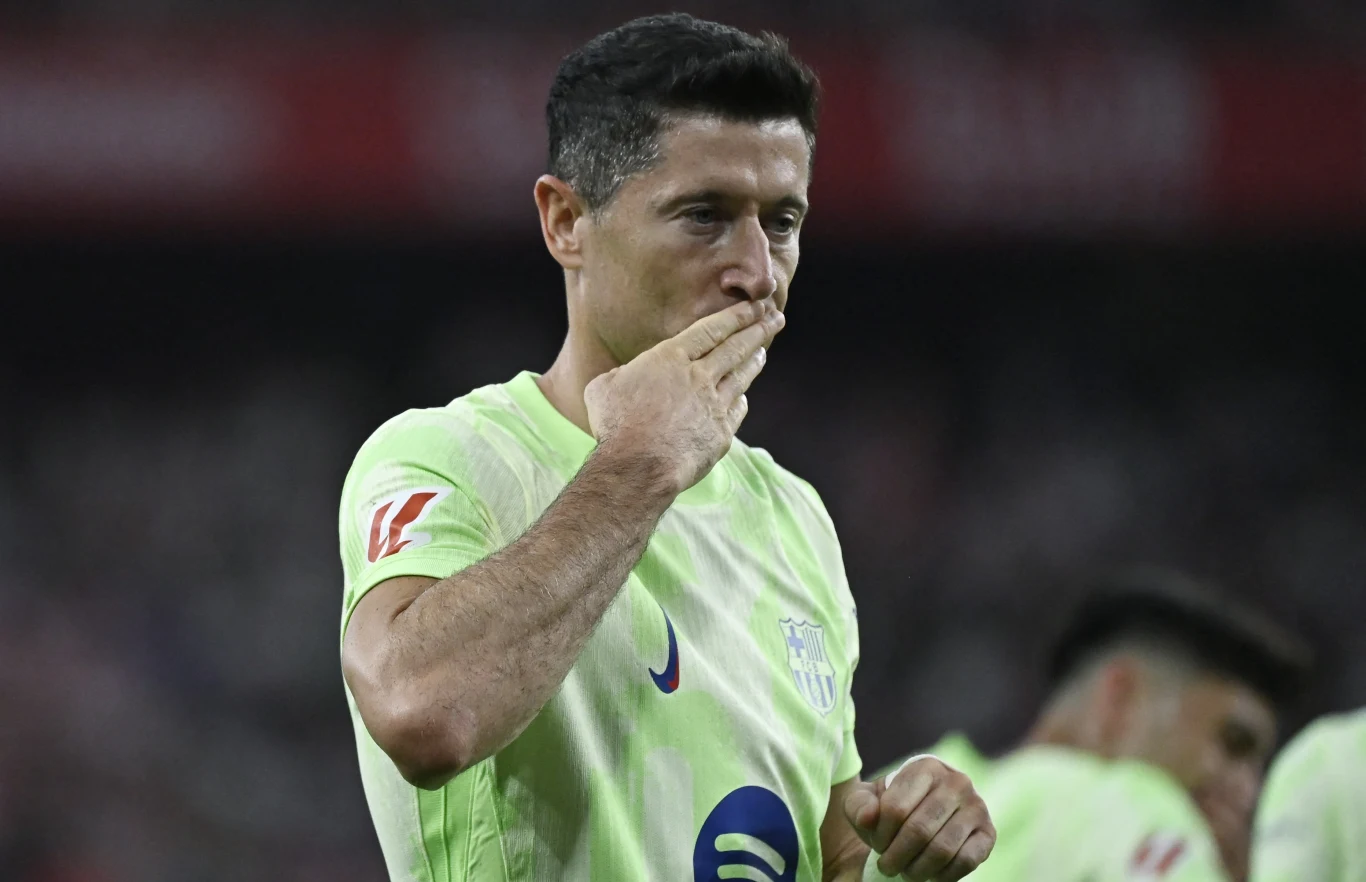 Robert Lewandowski Piłkarz w jasnozielonym stroju sportowym wykonuje gest pocałunku dłoni skierowany w stronę widowni na stadionie, w tle widoczni inni zawodnicy.