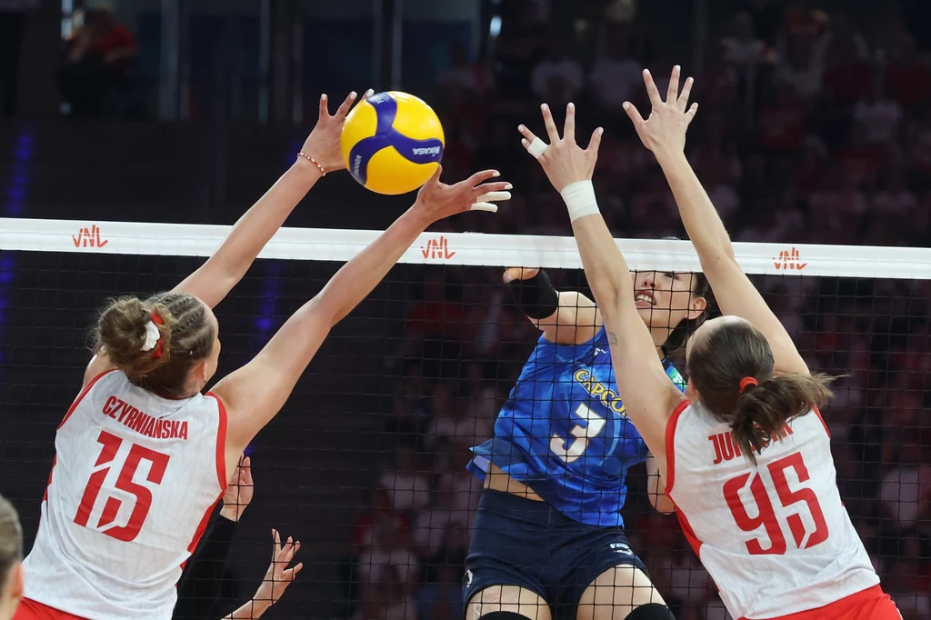 Magdalena Jurczyk (pierwsza z prawej) Dwie zawodniczki drużyny w białych strojach próbują zablokować uderzenie atakującej w niebieskim stroju na siatce, piłka siatkowa znajduje się tuż nad rękami zawodniczek, a w tle widoczni są kibice na trybunach.