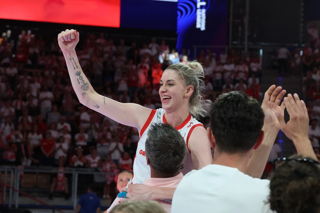 Magdalena Stysiak Zawodniczka drużyny siatkarskiej unosi w górę zaciśniętą pięść w geście radości, uśmiecha się szeroko i jest otoczona innymi osobami świętującymi sukces, a w tle widać trybuny pełne kibiców.