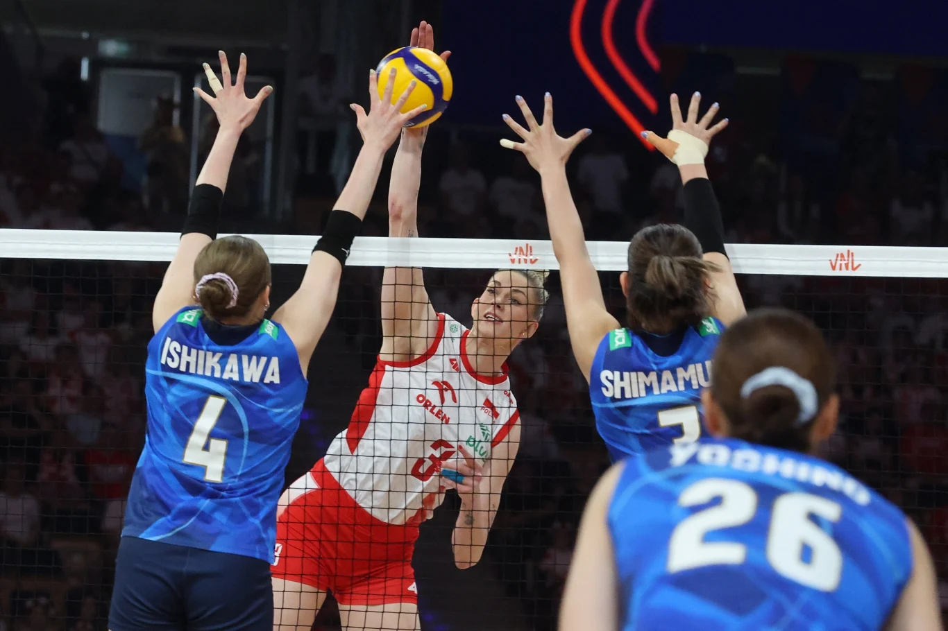 Magdalena Stysiak w ataku Zawodniczki dwóch drużyn siatkarskich rywalizują przy siatce podczas intensywnej akcji, środkowa w białym stroju atakuje piłkę, dwie przeciwne zawodniczki w niebieskich strojach próbują ją zablokować, widoczne koncentracja i sportowa rywalizacja.