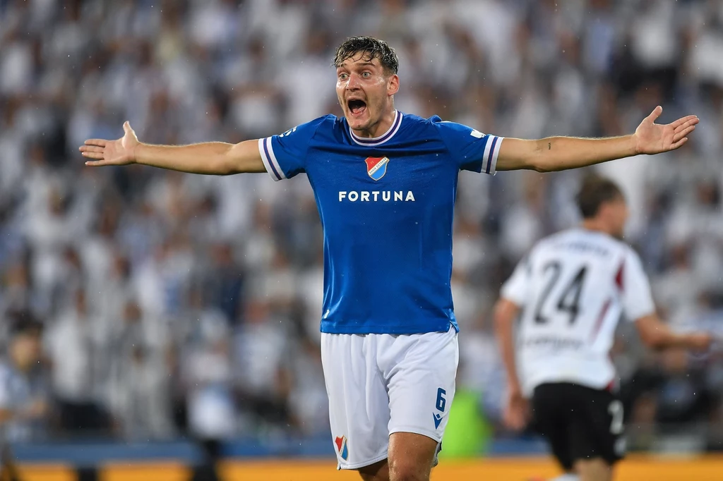 Karel Pojezny, środkowy defensor Banika Ostrawa Zawodnik w niebieskiej koszulce z numerem 6, o wyrazistej mimice, rozkłada ręce szeroko na stadionie podczas meczu piłkarskiego, w tle rozmyte sylwetki piłkarzy i kibiców.