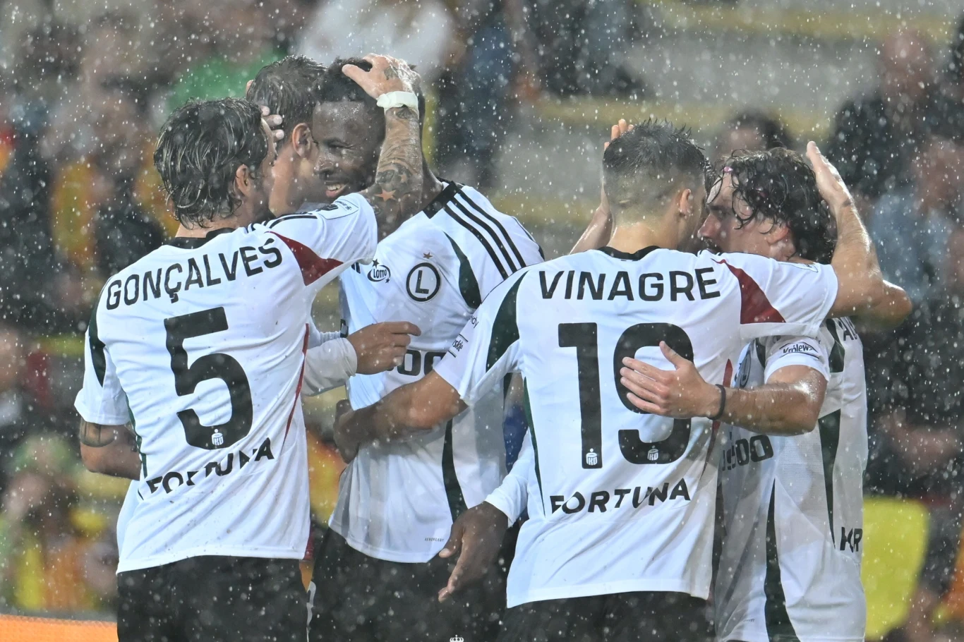 Grupa piłkarzy w białych koszulkach z nazwiskami świętuje zdobycie gola w deszczu, obejmując się i okazując radość, widoczne są nazwiska Gonçalves i Vinagre.