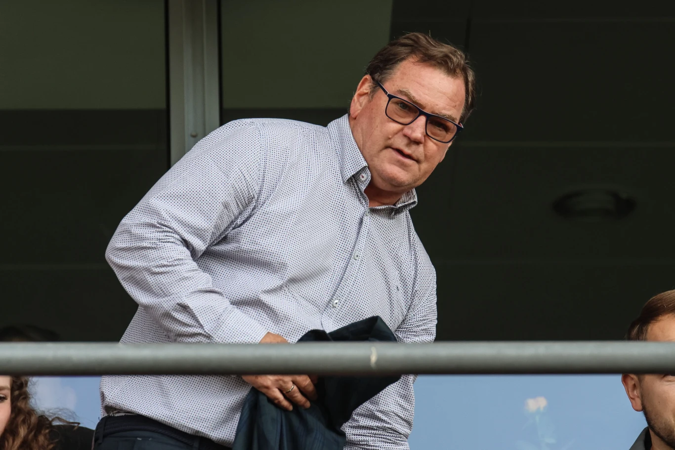 Jan Urban Mężczyzna w okularach i jasnej koszuli stoi przy balustradzie, trzyma marynarkę w rękach, obok widoczne fragmenty innych osób.