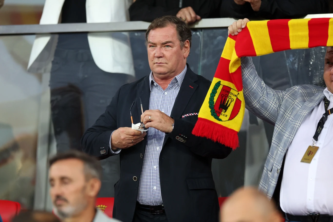 Jan Urban na meczu Korony Kielce z Legią Warszawa Mężczyzna w eleganckiej marynarce stojący na trybunach stadionu z poważnym wyrazem twarzy, obok niego osoba trzymająca żółto-czerwony szalik piłkarski.