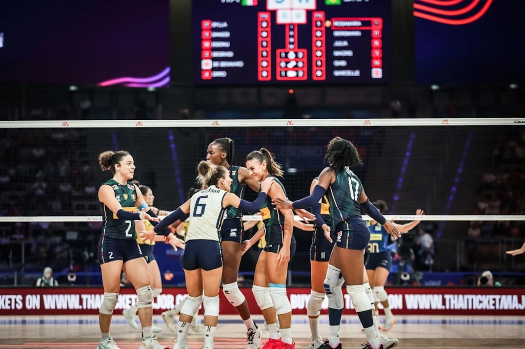 Siatkarki reprezentacji Włoch Grupa siatkarek świętuje zdobycie punktu podczas meczu na hali sportowej, wszystkie zawodniczki ubrane w reprezentacyjne stroje, w tle tablica wyników oraz publiczność.