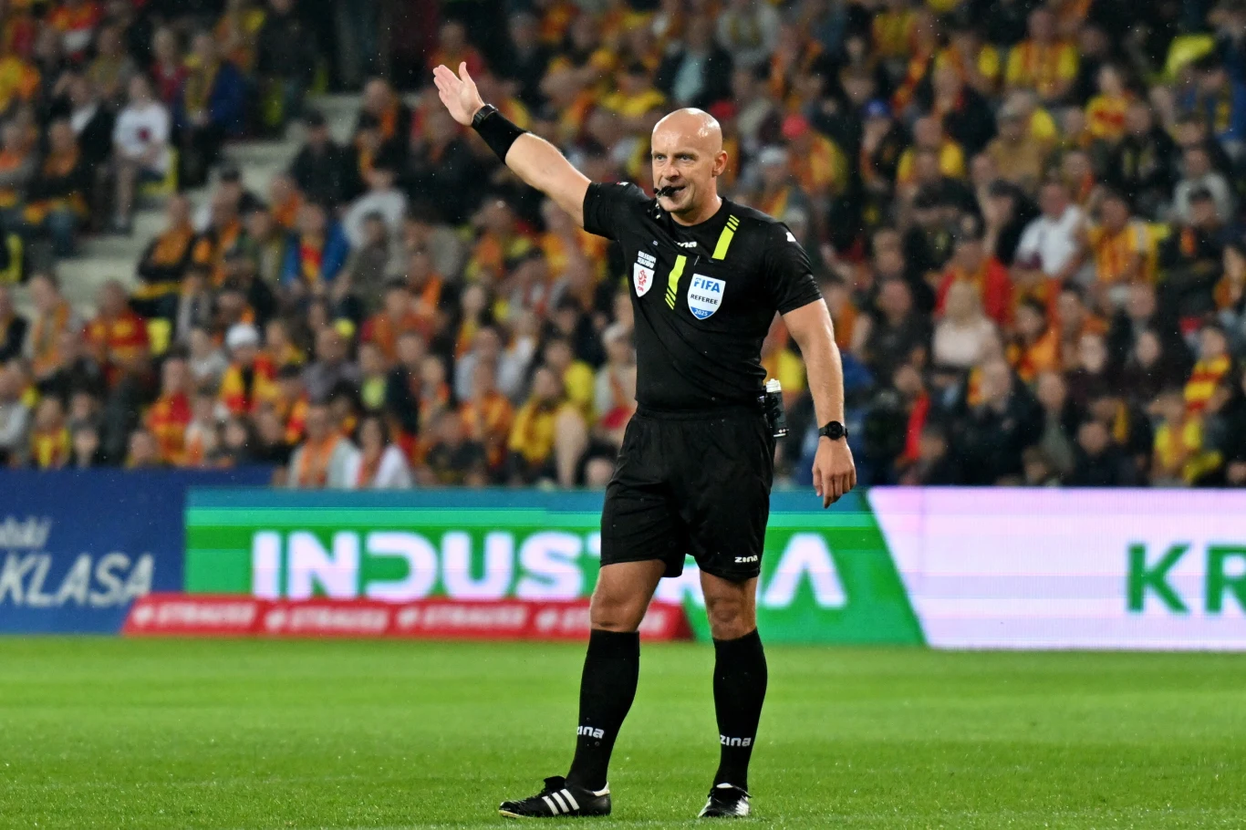 Sędzia piłkarski w czarnym stroju wskazuje kierunek na boisku, na tle trybun pełnych kibiców oraz reklamowych banerów.
