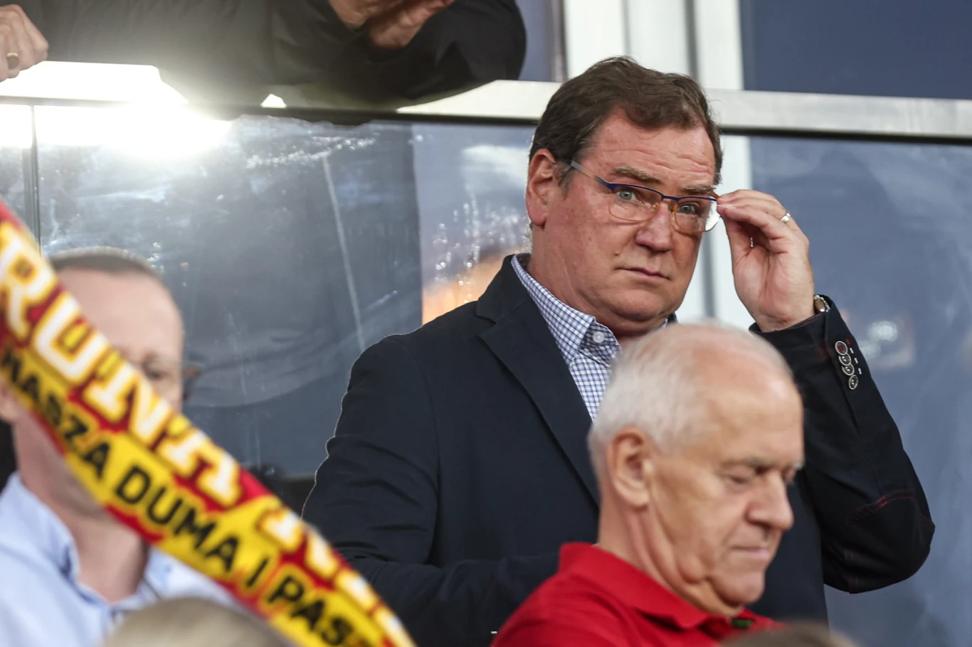 Mężczyzna w okularach, ubrany w granatową marynarkę i koszulę w kratkę, znajduje się na trybunach stadionu sportowego. W tle widać innych kibiców, a na pierwszym planie fragment szalika klubowego w czerwono-żółtych barwach.