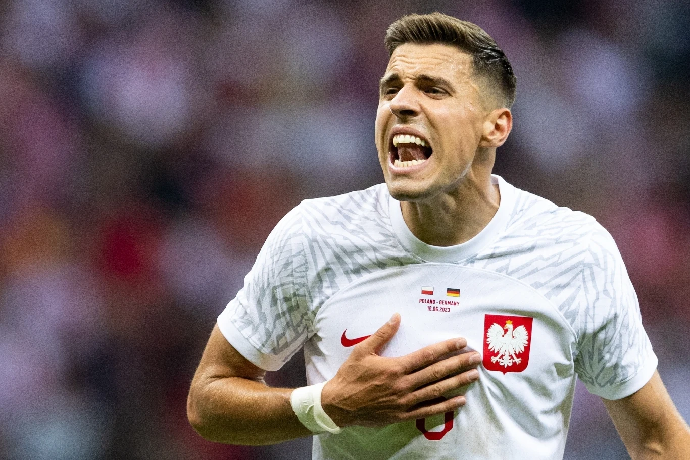 Jan Bednarek Piłkarz w białej koszulce z godłem Polski na piersi wyraża silne emocje podczas meczu, trzymając się za serce i głośno coś wykrzykując. W tle rozmazany tłum kibiców.