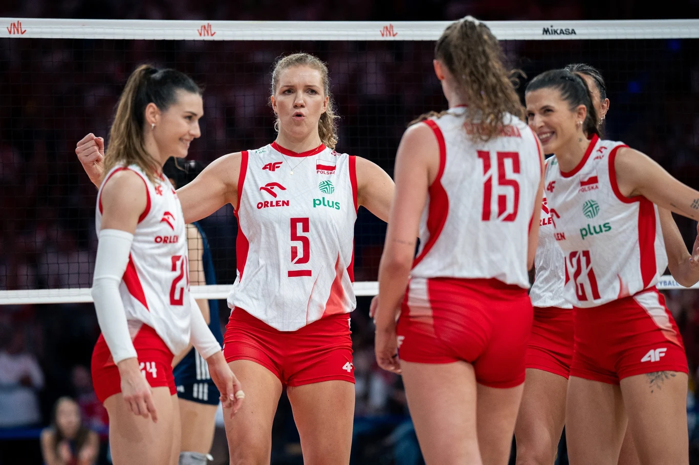 Agnieszka Korneluk (z numerem 5) w reprezentacji Polski Grupa zawodniczek drużyny siatkarskiej w biało-czerwonych strojach rozmawia na boisku podczas meczu, centralnie wyróżniona jedna z nich z uniesionymi ramionami, widoczne siatka oraz sportowa atmosfera.