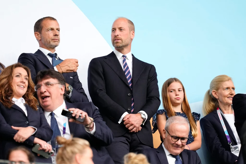 Grupa elegancko ubranych osób obserwuje wydarzenie sportowe z trybun, większość z nich ma poważny lub skupiony wyraz twarzy, w tle jasne, nowoczesne otoczenie stadionu.