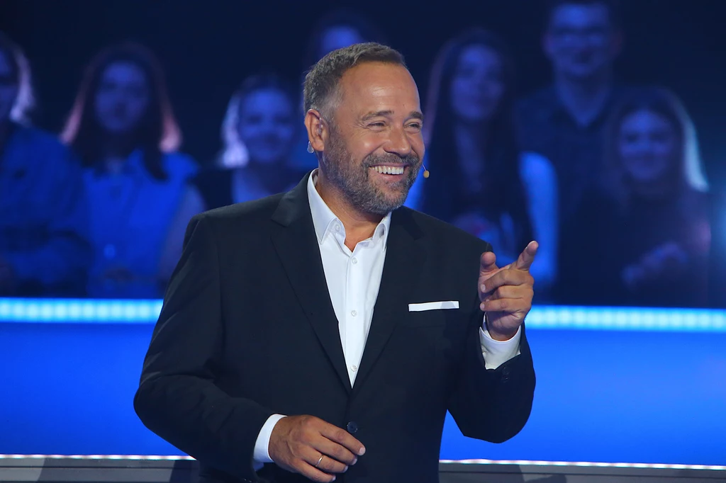Łukasz Nowicki nowym prowadzącym "The Voice Senior"! Mężczyzna w eleganckim, ciemnym garniturze z białą koszulą i mikrofonem na uchu uśmiecha się i wskazuje palcem, na drugim planie rozmyta publiczność siedząca na widowni