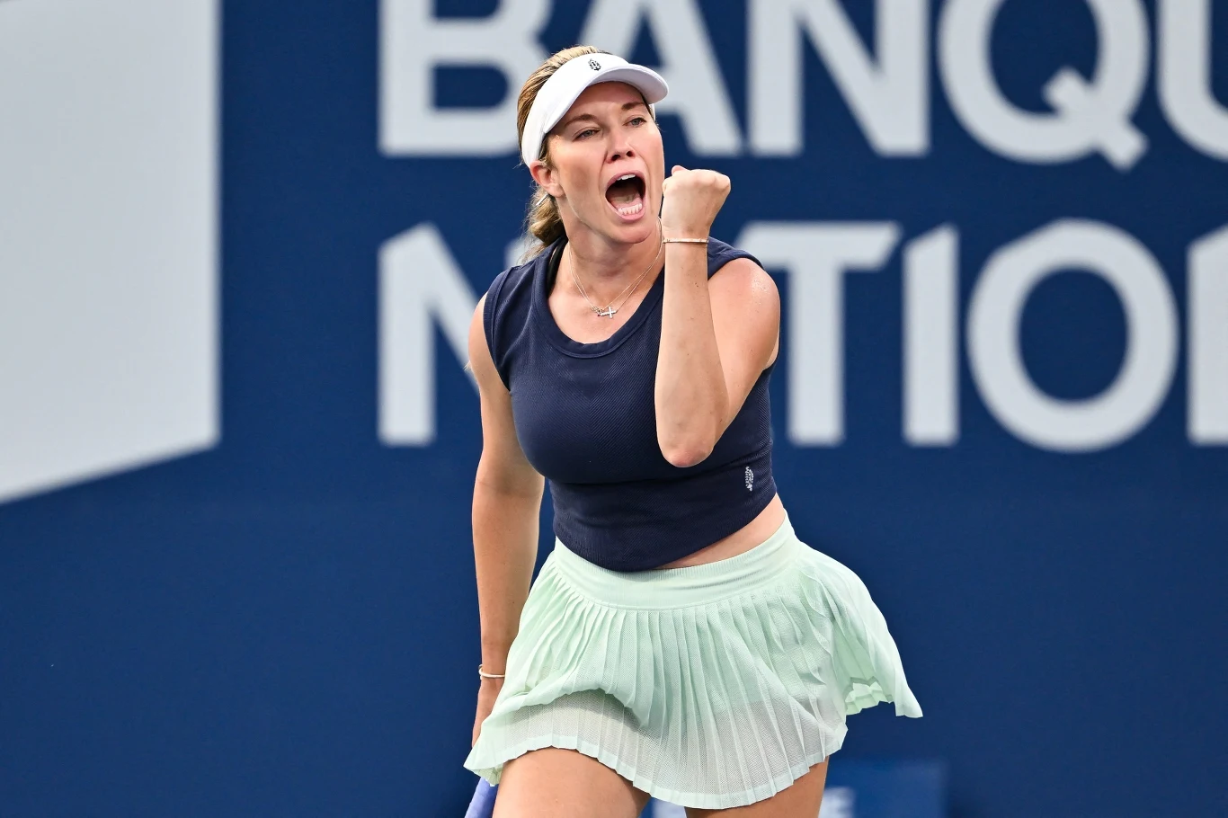 Tenisistka w sportowej odzieży z podniesioną pięścią wyraża emocje i determinację podczas meczu tenisowego na tle niebieskiego baneru reklamowego.