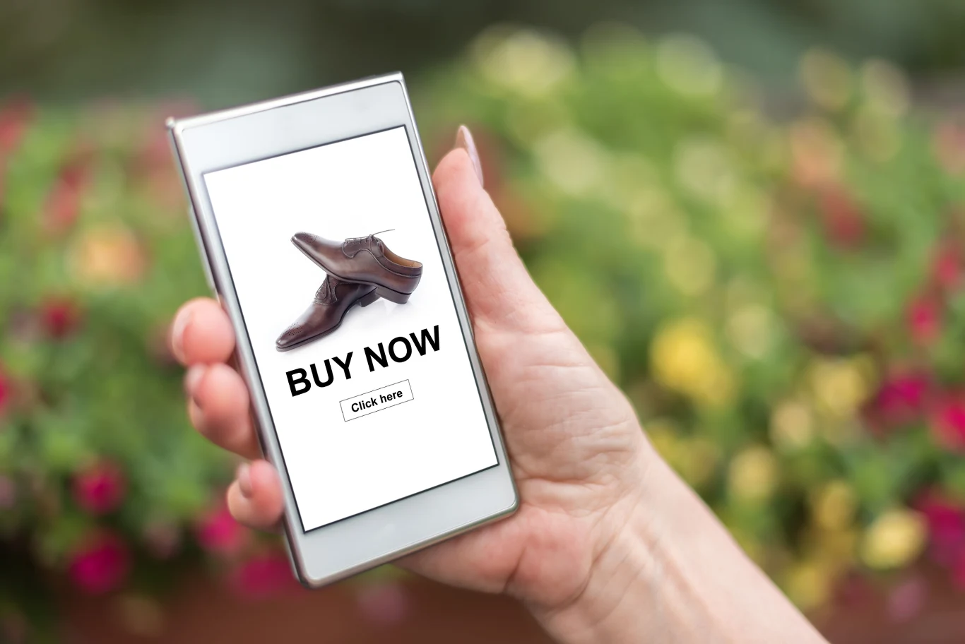 Dłoń trzymająca smartfona z widoczną stroną internetową prezentującą brązowy but i napis BUY NOW oraz przycisk Click here, tło rozmyte, zielono-różowe.