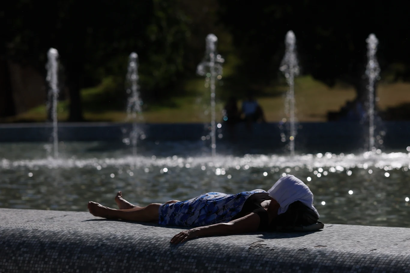 W miastach nawet cień staje się luksusem Osoba leżąca na brzegu fontanny w cieniu, z ręcznikiem lub tkaniną zakrywającą twarz, w tle tryskająca woda z kilku strumieni fontanny, otoczenie zielone, letnia atmosfera.
