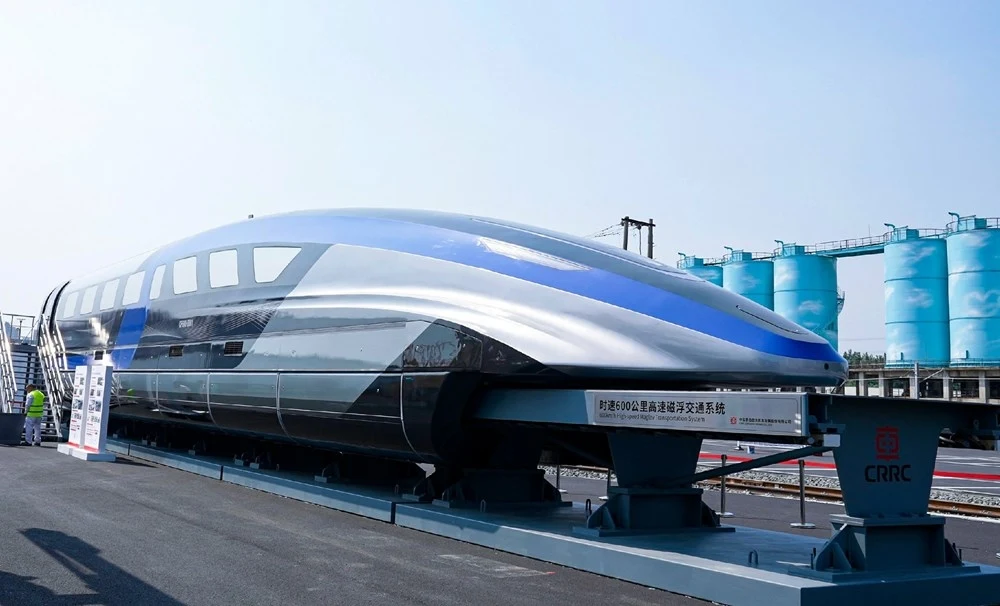 Chińskie Koleje CRRC prezentują pociąg maglev dla prędkości 600 km/h. Nowoczesny pociąg typu maglev o aerodynamicznym, metalicznym nadwoziu z domieszką niebieskiego ustawiony na torze pokazowym, w tle cylindryczne zbiorniki przemysłowe w jasnoniebieskim kolorze.
