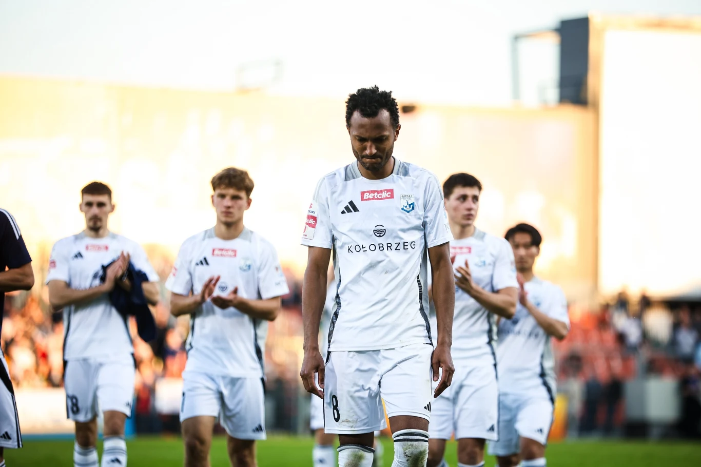 Zawodnikom Kotwicy Kołobrzeg nie udało się utrzymać w I lidze. Spadek przypieczętował upadek klubu Zawodnicy drużyny piłkarskiej w białych strojach schodzą z boiska, na twarzach mają poważne wyrazy, w tle trybuny pełne kibiców.