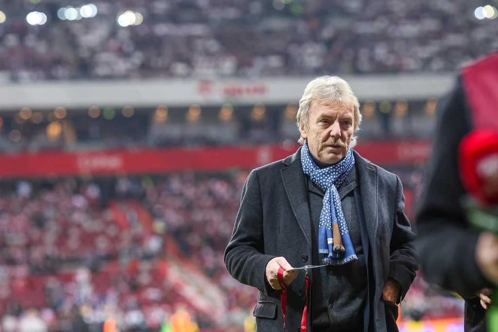 Zbigniew Boniek Starszy mężczyzna ubrany elegancko w ciemny płaszcz i niebieski szalik, stoi na tle stadionu wypełnionego widzami podczas wydarzenia sportowego.