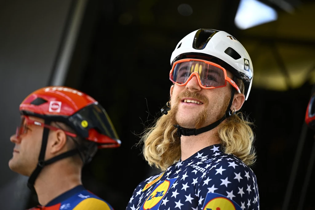 Quinn Simmons Dwóch mężczyzn w kaskach rowerowych, ubranych w profesjonalne stroje kolarskie, jeden z nich z długimi blond włosami i uśmiechem, obaj mają okulary sportowe i wydają się znajdować przed rozpoczęciem wyścigu.