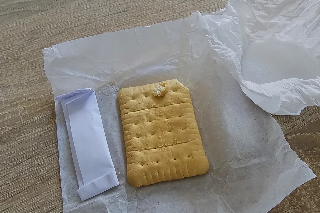 Duży suchar leży na kawałku papieru, obok znajduje się zwinięty w papier prostokątny przedmiot, najprawdopodobniej opakowanie suchara.