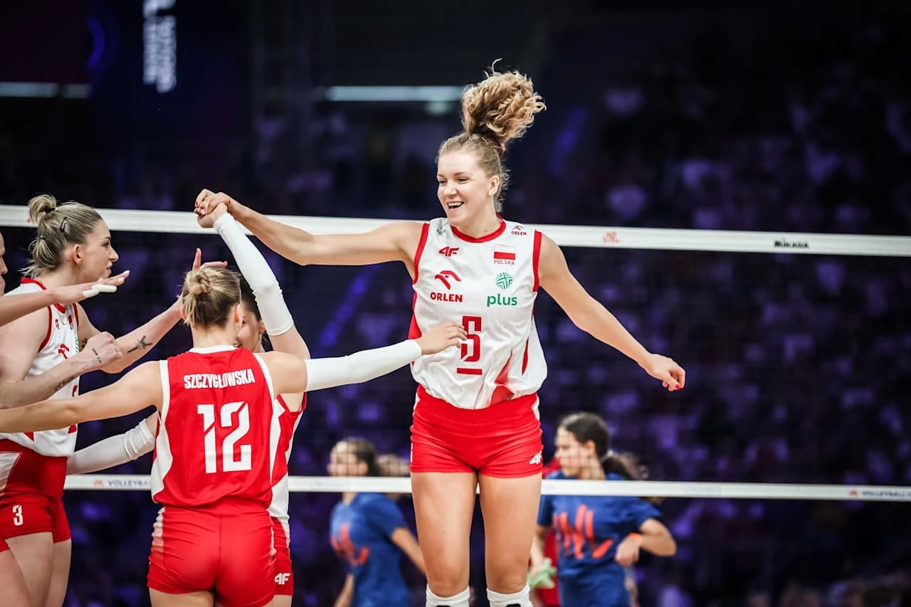 Siatkarki reprezentacji Polski Polska siatkarka w biało-czerwonym stroju cieszy się po zdobytym punkcie, unosząc rękę i podskakując, wokół niej są koleżanki z zespołu na tle siatki i zawodniczek drużyny przeciwnej.