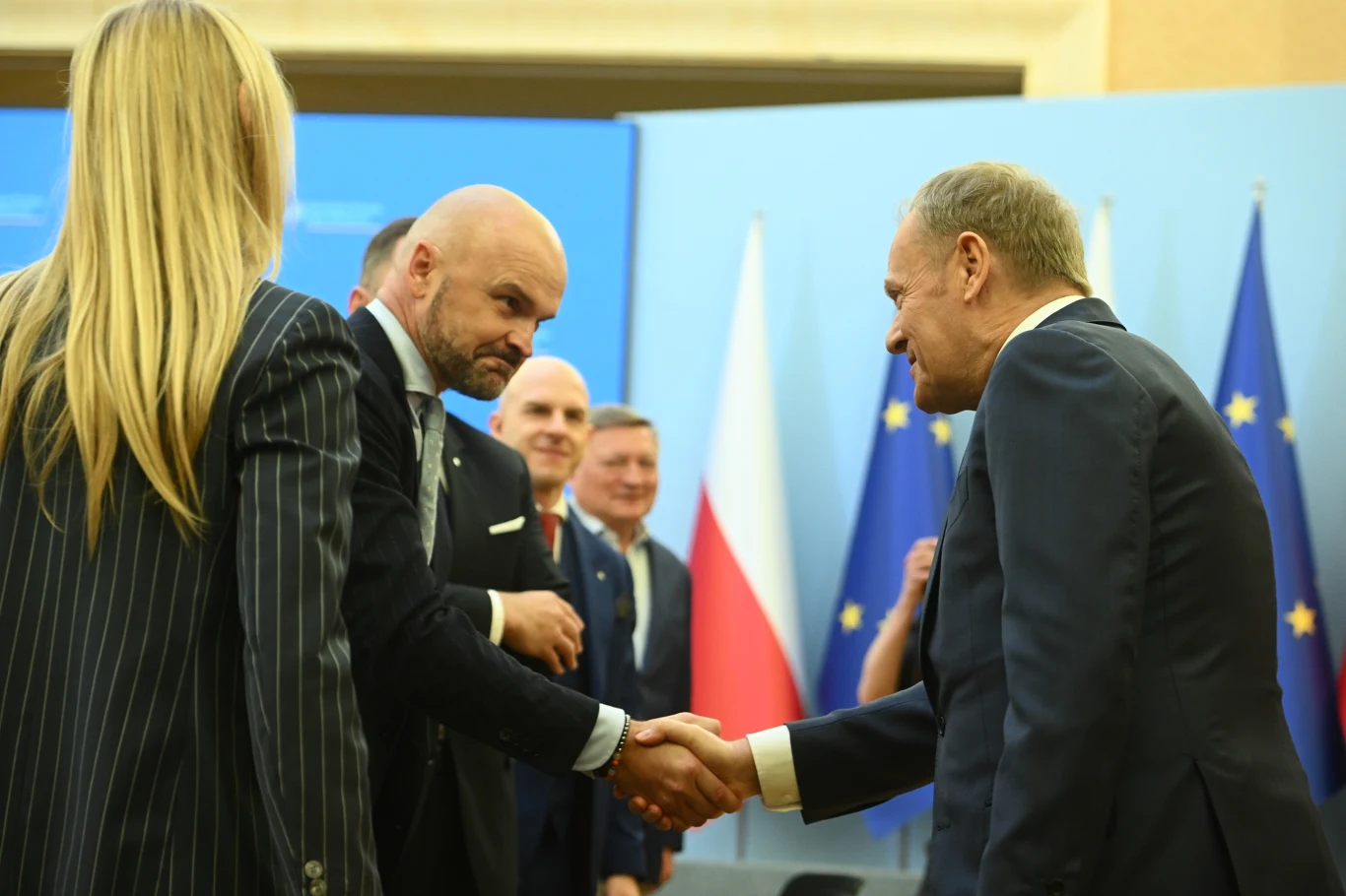 Rafał Brzoska i Donald Tusk Grupa polityków podczas oficjalnego spotkania, mężczyźni w garniturach ściskają sobie dłonie na tle flag Polski oraz Unii Europejskiej.