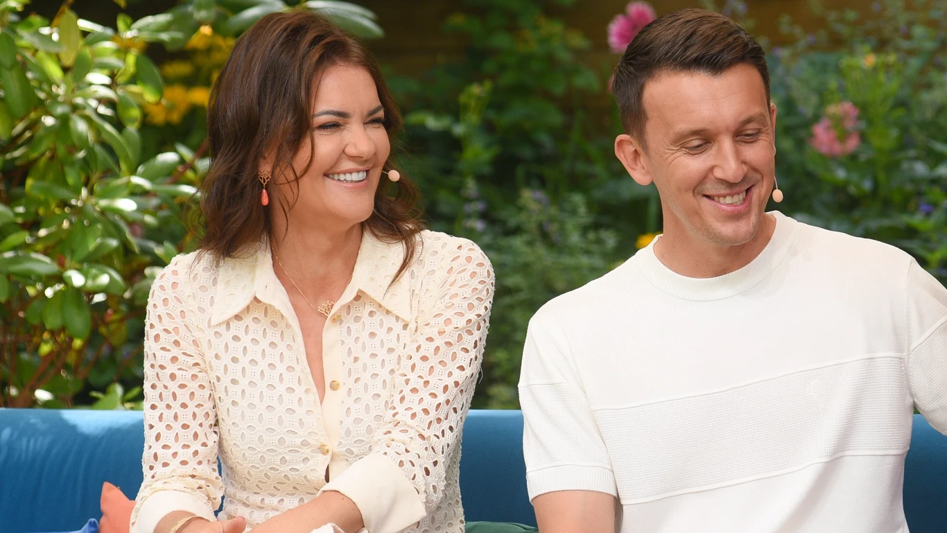 Agnieszka Radwańska i Dawid Celt Dwie osoby siedzące obok siebie na tle zielonych roślin, z uśmiechami na twarzach, oboje mają przypięte mikrofony do ubrania, panuje przyjazna atmosfera.