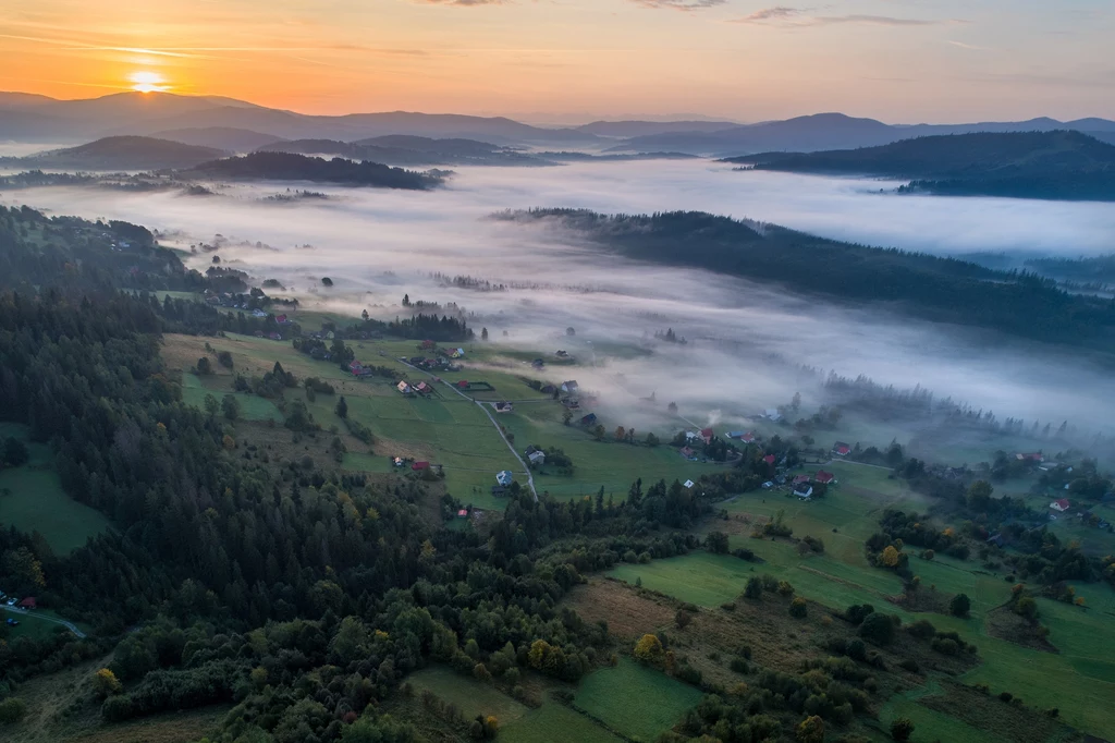 Ochodzita oferuje taką panoramę Poranna mgła unosząca się nad malowniczymi polami, lasami oraz niewielkimi domami w dolinie, przy łagodnym świetle wschodzącego słońca na tle górskich pasm.