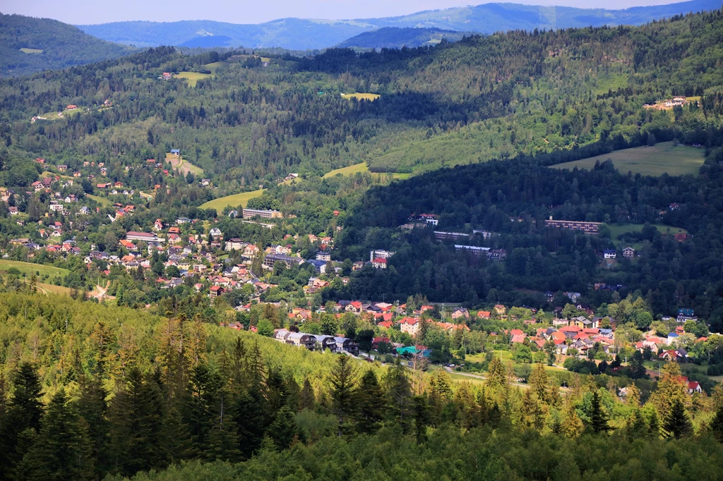 Widok na Wisłę - miejscowość, która może być początkiem niejednej górskiej wędrówki Panorama górskiej miejscowości otoczonej lasami, rozległa zabudowa domków jednorodzinnych w dolinie, w tle wzniesienia porośnięte zielonymi drzewami i niewielkie pola.