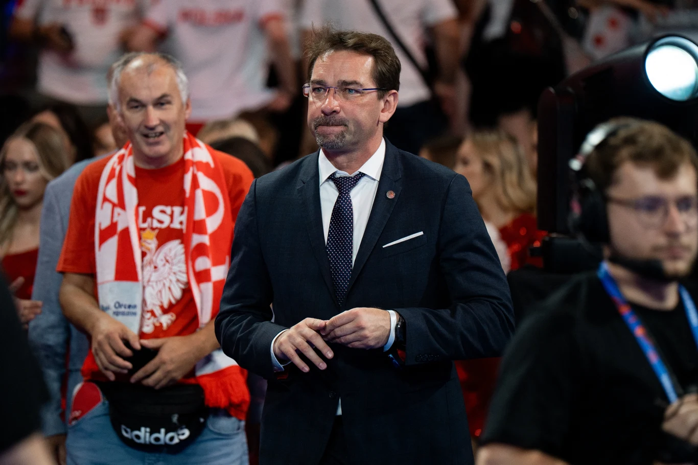 Mężczyzna w garniturze i okularach znajduje się na pierwszym planie w hali sportowej, za nim widoczny tłum kibiców ubranych w biało-czerwone stroje i szaliki z orłem oraz napisem 'Polska'.