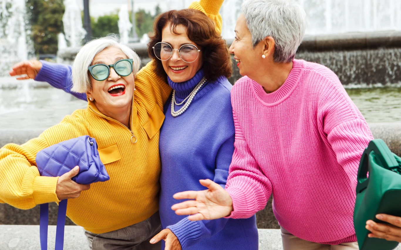 Trzy radosne starsze kobiety w kolorowych swetrach stoją przy fontannie, obejmują się i uśmiechają, tworząc pozytywną i przyjazną atmosferę.