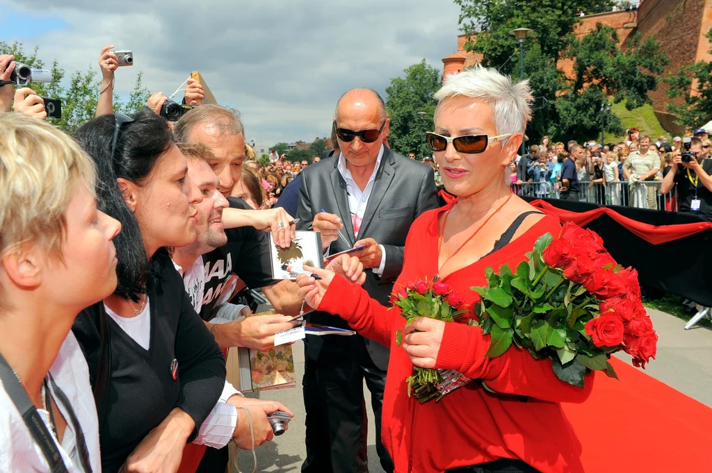 Marek Jackowski i Kora (Maanam) w 2009 r. Kobieta z krótkimi, jasnymi włosami i w okularach przeciwsłonecznych ubrana w czerwoną bluzę trzyma bukiet czerwonych róż i rozdaje autografy grupie fanów. W tle liczna publiczność, fotografowie oraz osoby dokumentujące wydarzenie.
