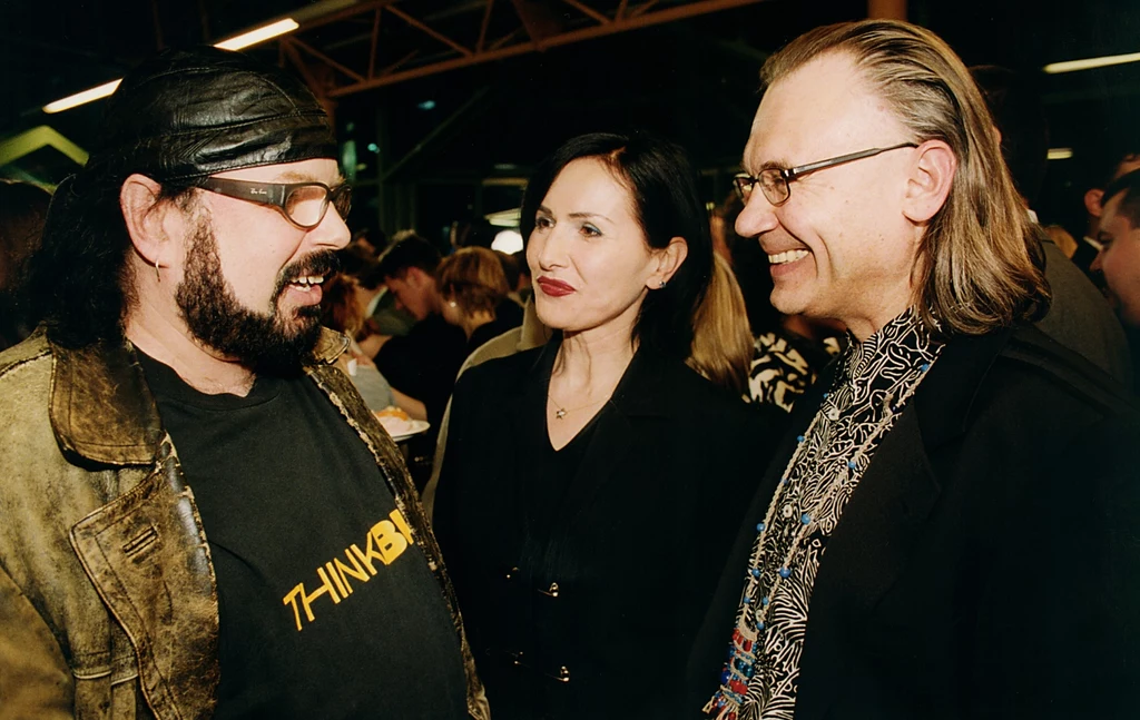 Zbigniew Hołdys, Kora i Kamil Sipowicz w 2001 r. Trzy osoby rozmawiają w tłumie podczas wydarzenia towarzyskiego, dwie z nich się uśmiechają, a mężczyzna po lewej stronie nosi czarną koszulkę z napisem i skórzaną kurtkę.