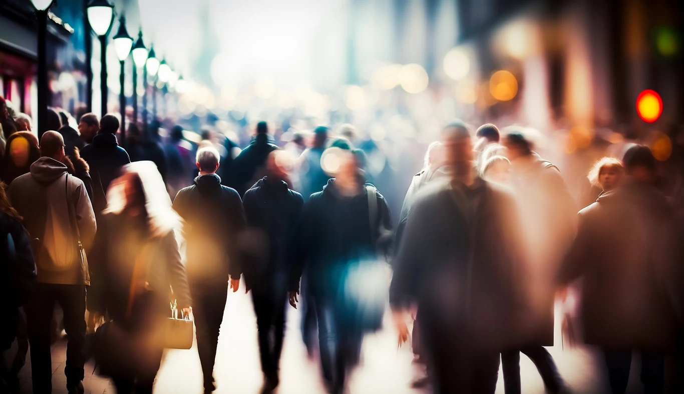 Miasto w biegu. Piesi przyspieszyli i rzadziej przystają w miejscach publicznych Tłum ludzi idących ulicą w dużym mieście, rozmyte sylwetki nadają scenie dynamiczny i nieco anonimowy charakter, widoczne latarnie i miejskie światła tworzą atmosferyczny nastrój.