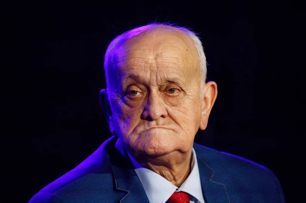 Starszy mężczyzna z poważnym wyrazem twarzy, ubrany w granatową marynarkę, białą koszulę i czerwoną krawatę, na ciemnym tle.