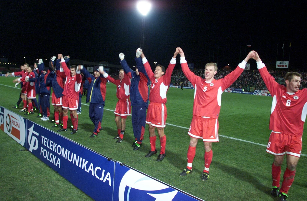 Grupa piłkarzy w czerwonych strojach trzymających się za ręce i unoszących je w geście triumfu na murawie stadionu, w tle jasne światła reflektorów oraz kibice na trybunach.