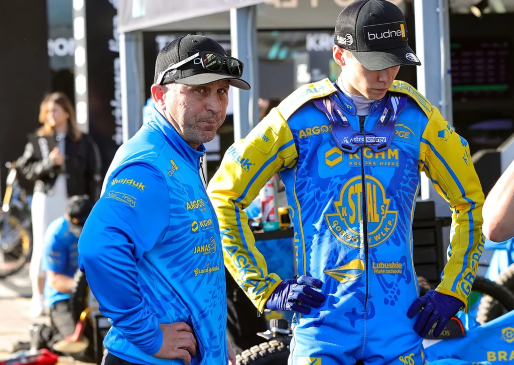 Oskar Paluch z tatą Piotrem. Dwóch mężczyzn w niebiesko-żółtych strojach drużyny sportowej stoi obok siebie, jeden patrzy w stronę aparatu, drugi ze spuszczoną głową z założonymi rękami, w tle widoczne wyposażenie i członkowie drużyny.