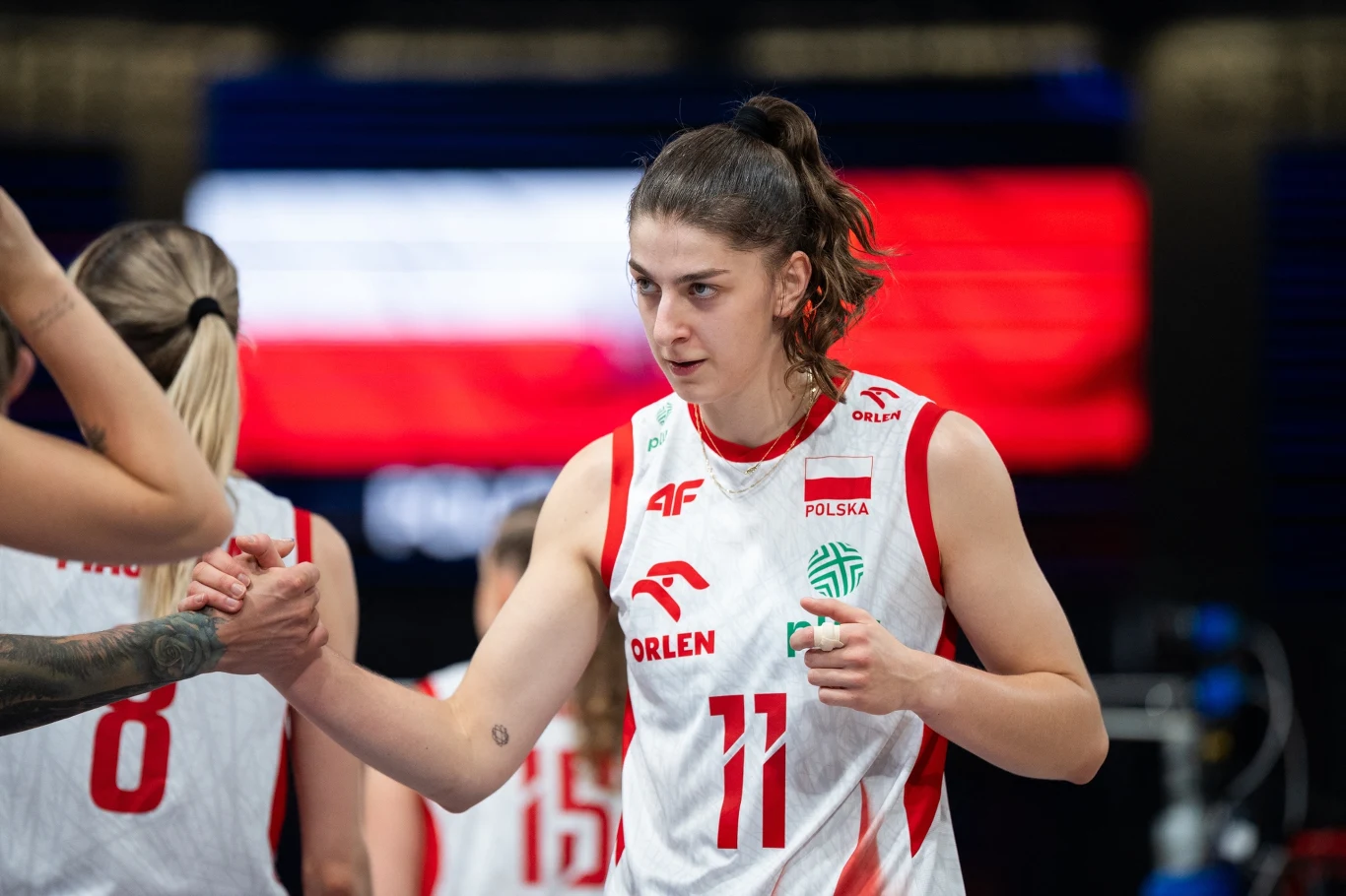 Zawodniczka polskiej drużyny koszykarskiej z numerem 11 przybija piątkę z inną zawodniczką, obie ubrane w oficjalne stroje sportowe, w tle rozmyty fragment flagi Polski i inni członkowie zespołu.