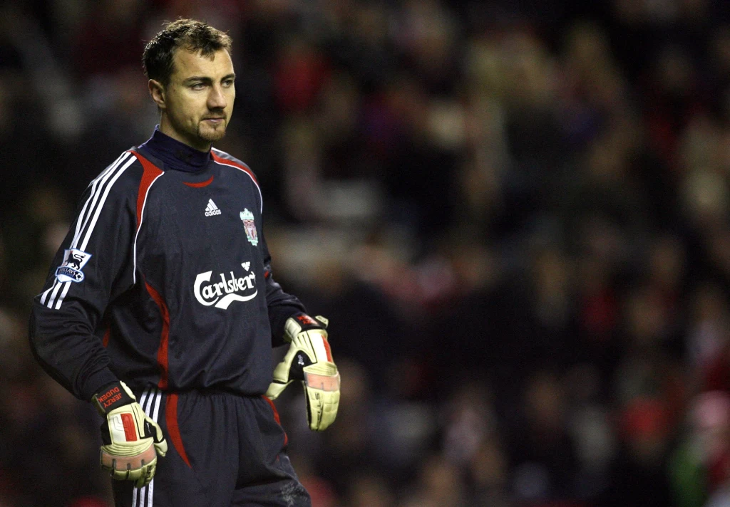 Jerzy Dudek Bramkarz w ciemnym stroju sportowym Liverpool FC stoi na murawie podczas meczu piłkarskiego, trzymając rękawice, w tle widoczna rozmazana publiczność.