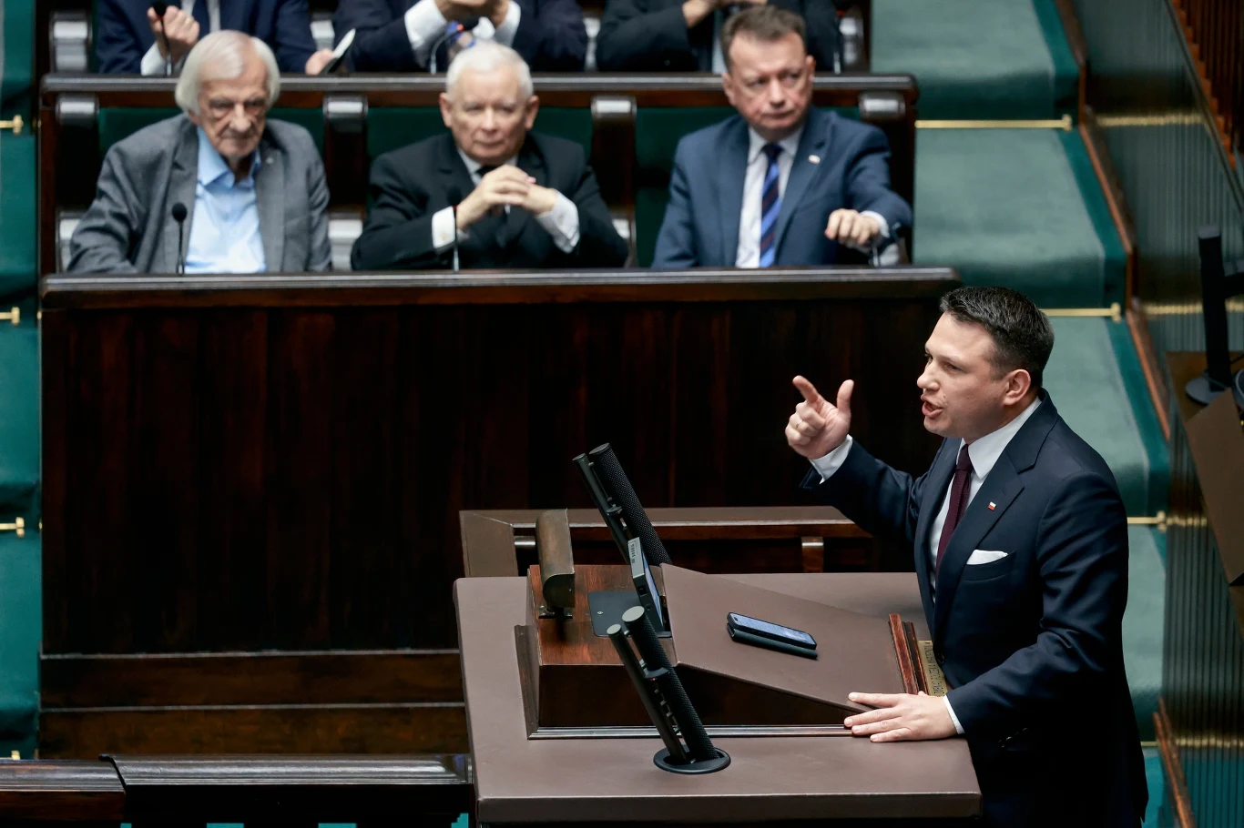 Polityk przemawia przy mównicy w sali sejmowej, wokół znajduje się kilku innych polityków słuchających wystąpienia, tło stanowią ławki poselskie.