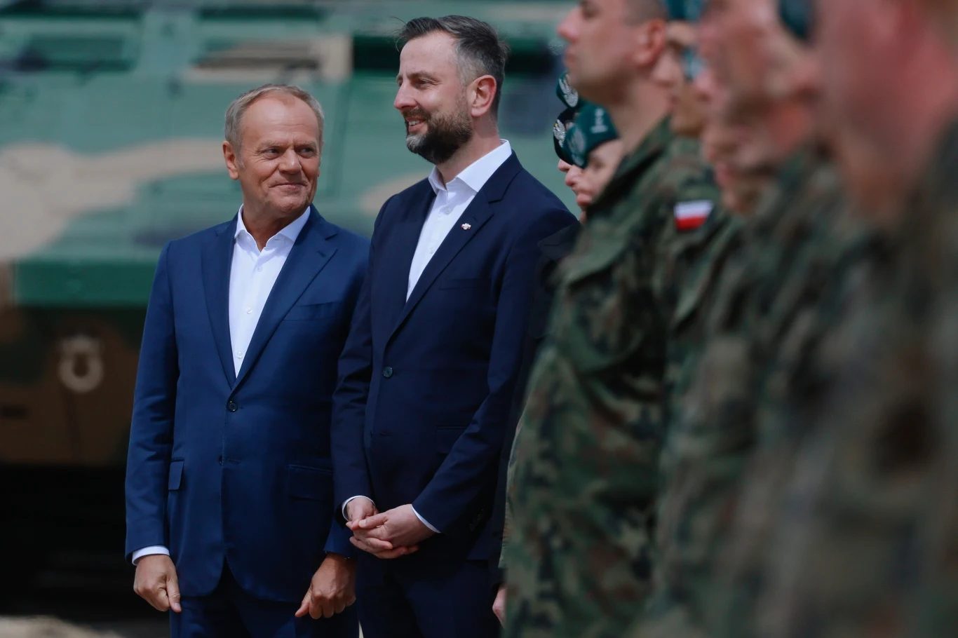 Donald Tusk zawsze cenił współpracę z Władysławem Kosiniakiem-Kamyszem Dwóch mężczyzn w garniturach stoi obok siebie na tle zamazanego pojazdu wojskowego, po prawej stronie ustawieni w szeregu żołnierze w mundurach.