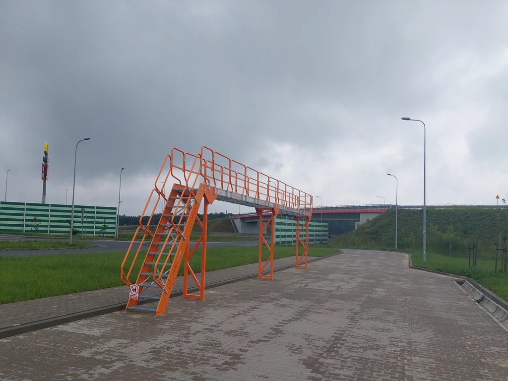 Pomarańczowa metalowa konstrukcja z podestem i poręczami ustawiona na brukowanej drodze, otoczona zielenią, z widocznym wiaduktem i ekranami akustycznymi w tle pod pochmurnym niebem.