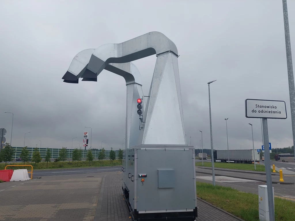 Maszyna do odśnieżania pojazdów ustawiona przy jezdni, z metalowymi ramionami i sygnalizatorem świetlnym, w tle parking oraz znak informujący o stanowisku do odśnieżania.