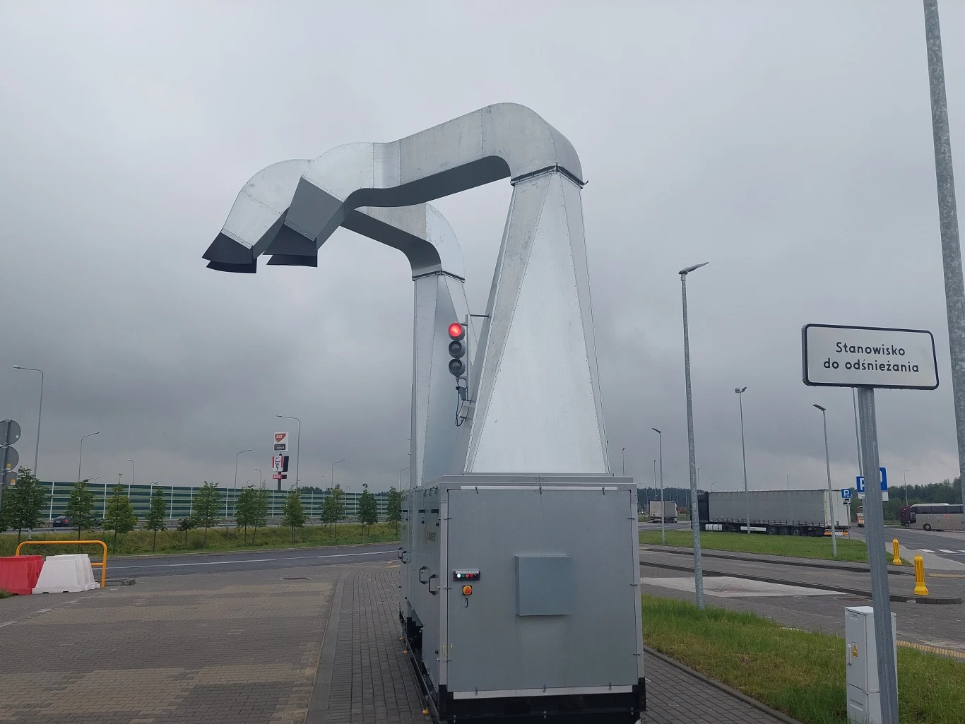 Maszyna do odśnieżania pojazdów ustawiona przy jezdni, z metalowymi ramionami i sygnalizatorem świetlnym, w tle parking oraz znak informujący o stanowisku do odśnieżania.